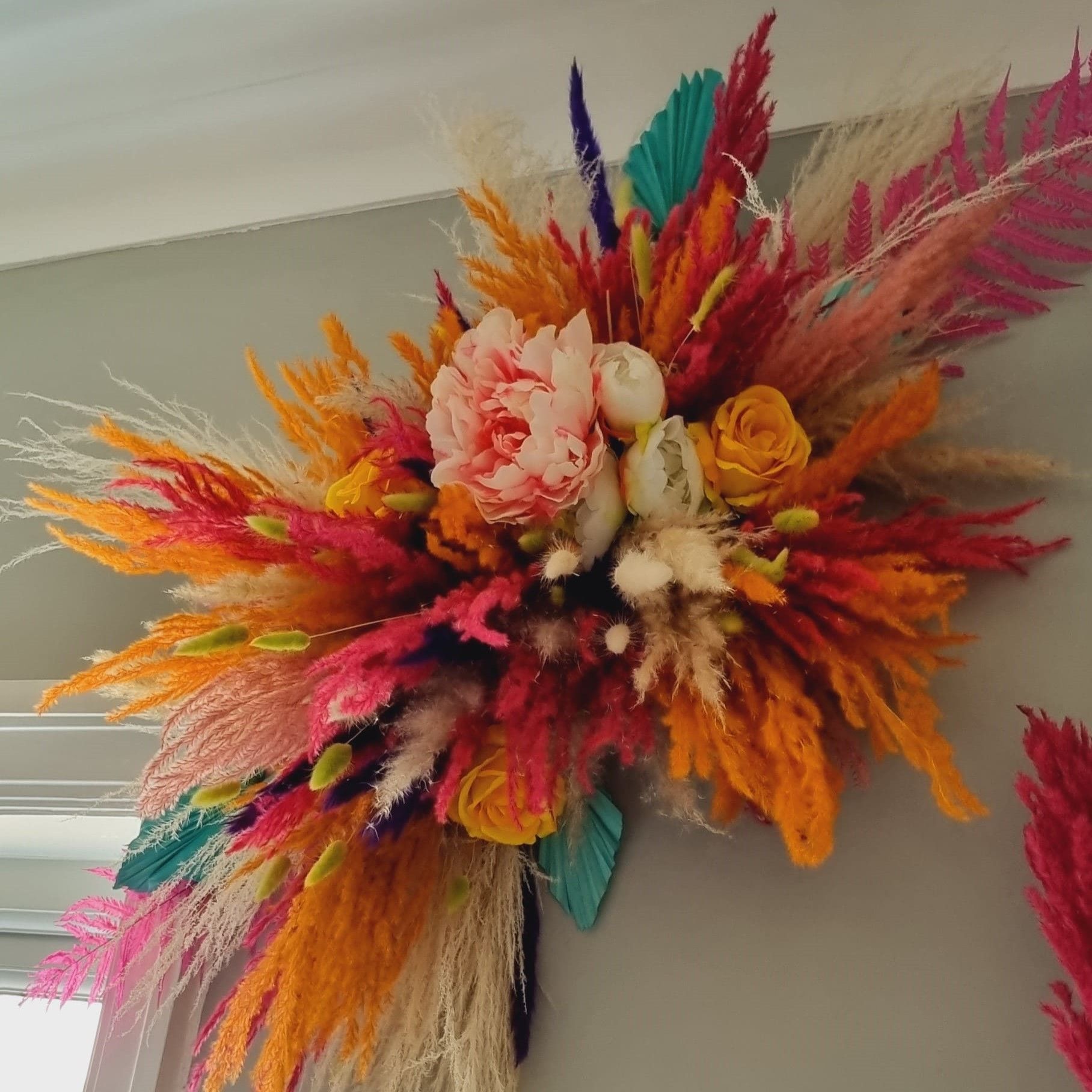 bright coloured dried pampas grass wall decor, multicoloured pampas wall art, cerise pink pampas grass, orange pampas grass, purple mini pampas, blue speared palms with artifical peony and yellow roses.