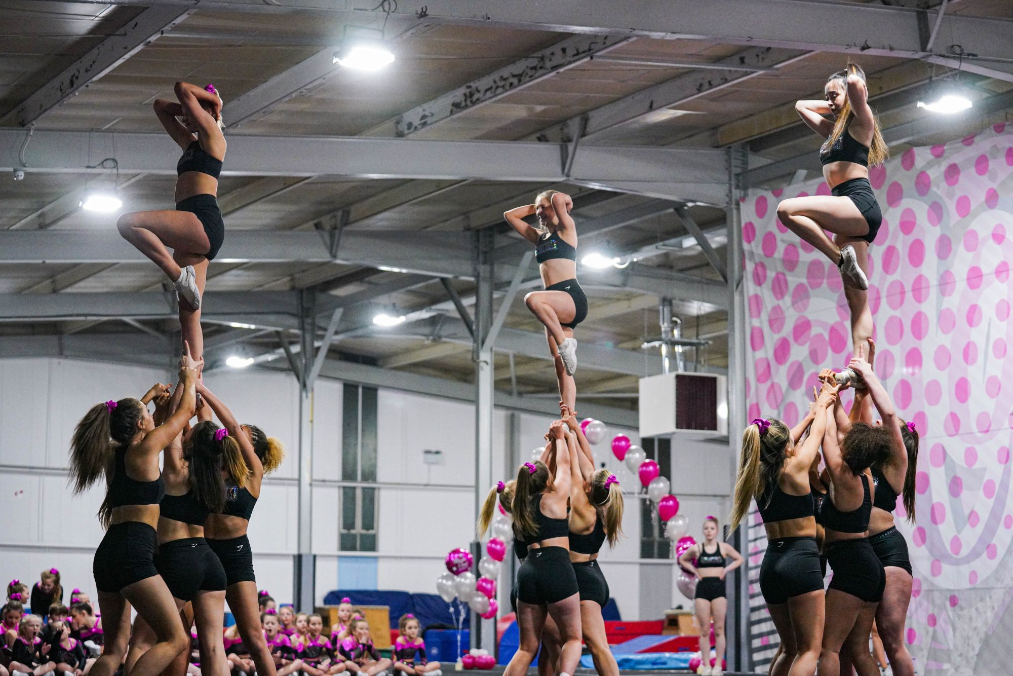 Three groups of girls, each supporting a girl who they are holding above them and who is balancing on one leg.