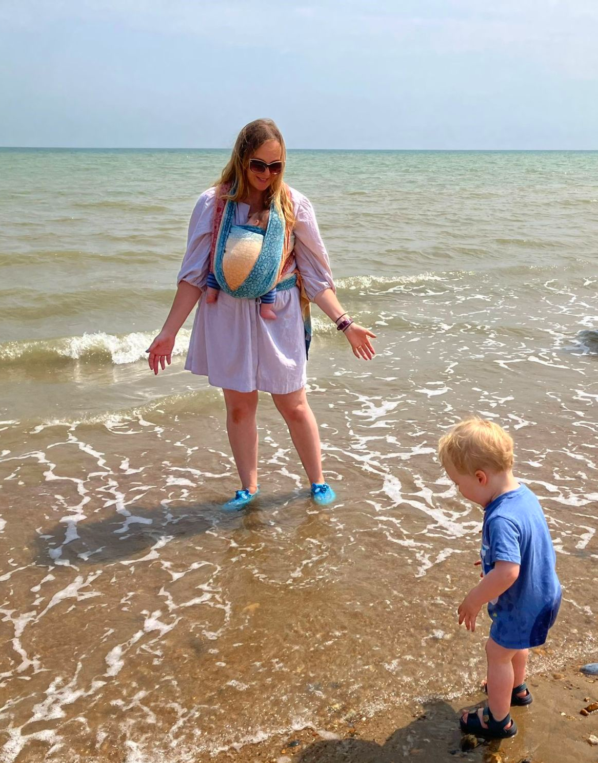 the hypnobirthing teacher with her two children - she is standing in shallow water at the sea with the baby wrapped in a colourful sling on her chest, while her toddler stands in front of her, looking for pebbles in the sea