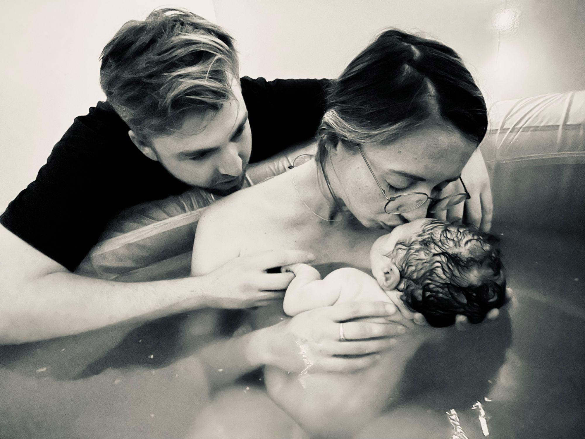 homebirth a new mum kisses her newborn, while her partners hugs them both in the birth pool
