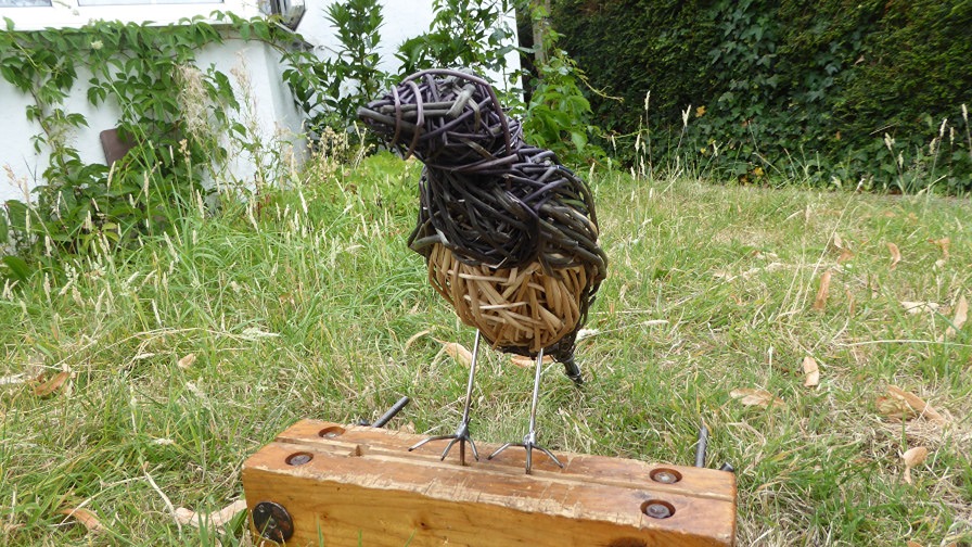 magpie sculpture made from willow , black and creme standing on woooden base places on grass 