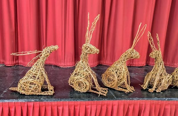 willow hares  sitting on a stage in a hall m with a red curtain behind them 