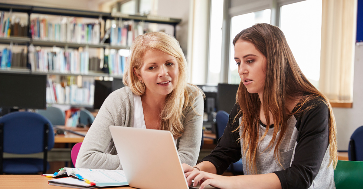 teacher supporting a student with work on a laptop in a library setting