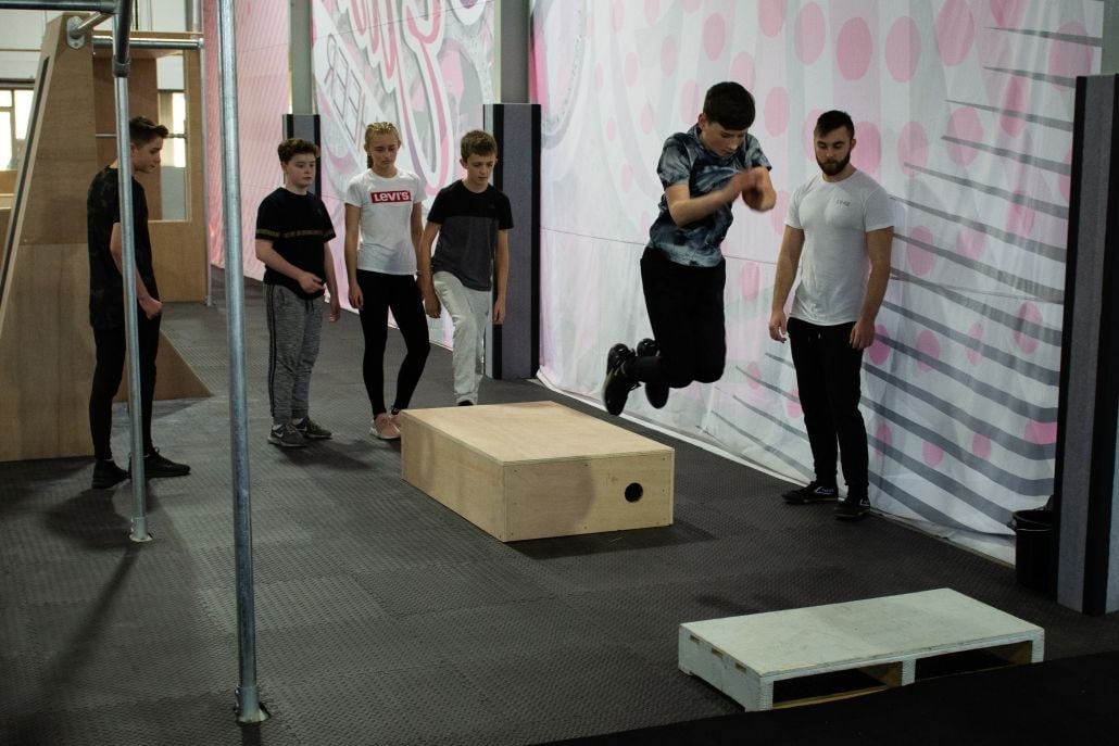 A boy jumping between two blocks as others wait behind him and the coach looks on.