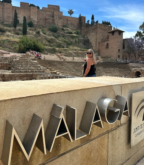 Modern M&Atilde;&iexcl;laga city sign with the Roman Theatre and Alcazaba fortress blurred in the background.
