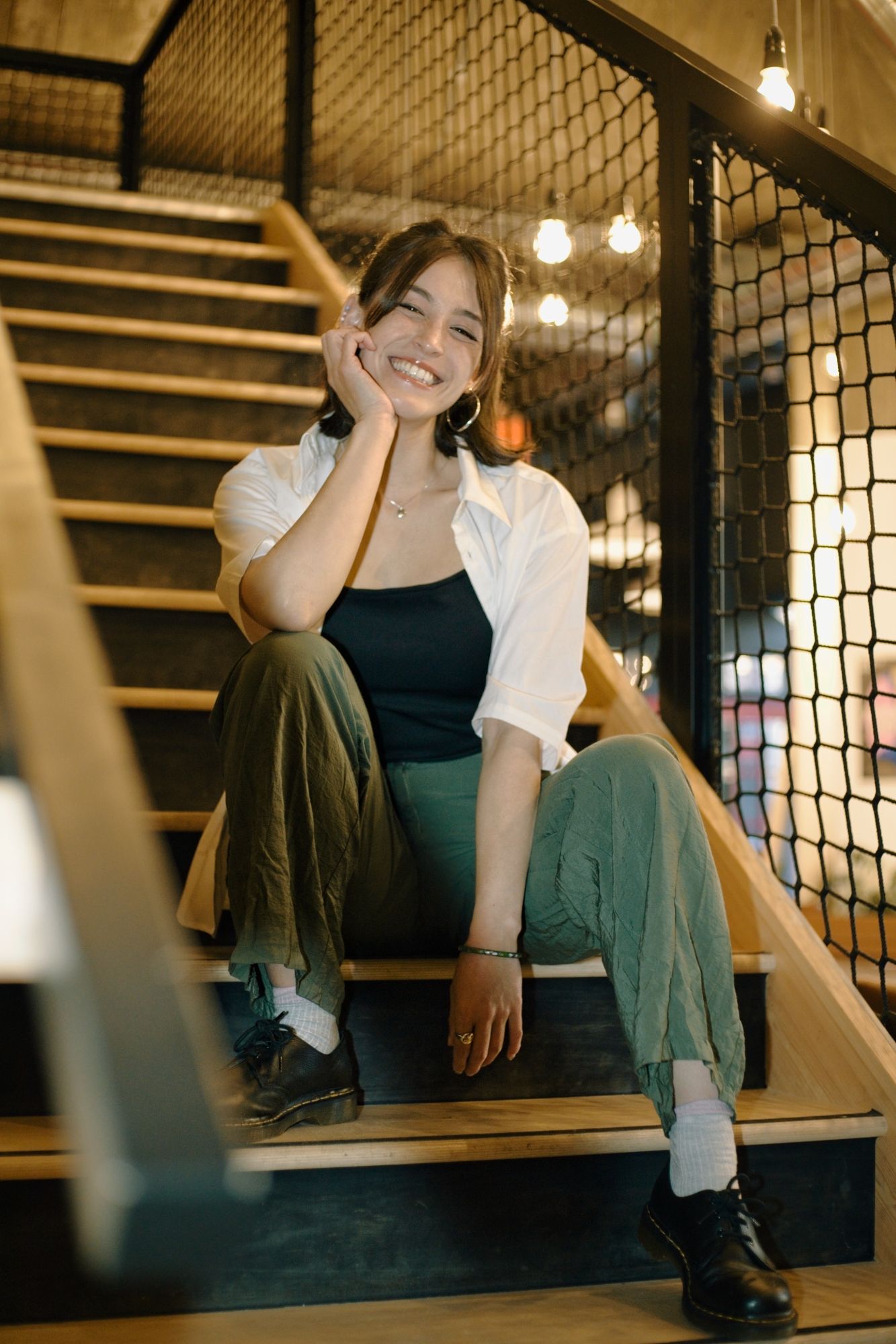 A Vietnamese-British woman wearing green trousers and a white blouse sits on a staircase, smiling at the camera. Her head leans on her right hand and she looks relaxed.