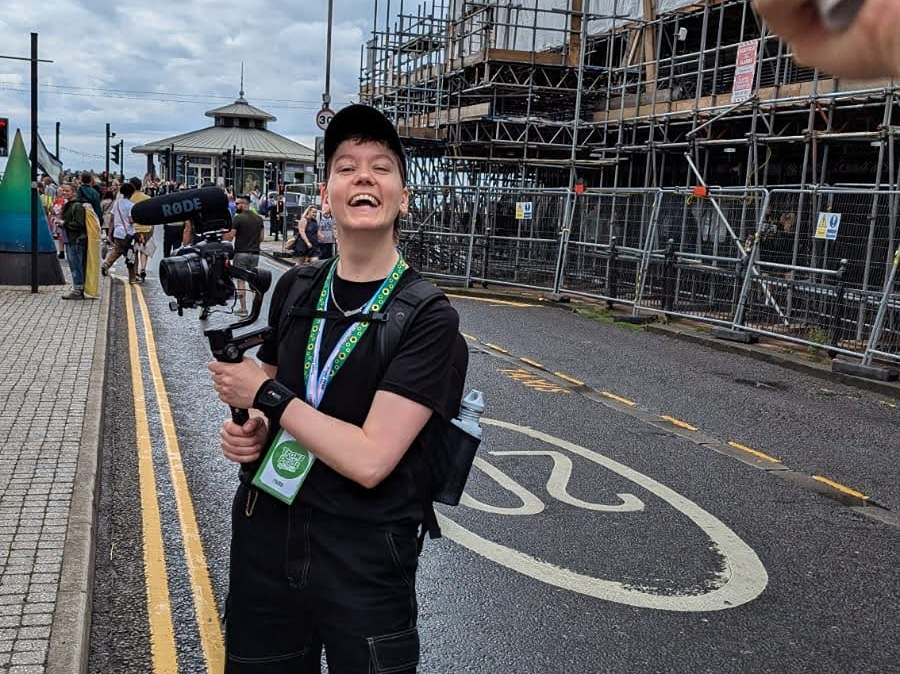 Roxy films a march for Trans Pride Brighton with a big smile on their face. They're holding a camera and microphone on a stabiliser. They wear all black clothes including a cap, and have two lanyards around their neck, one for Trans Pride Brighton and one sunflower lanyard.