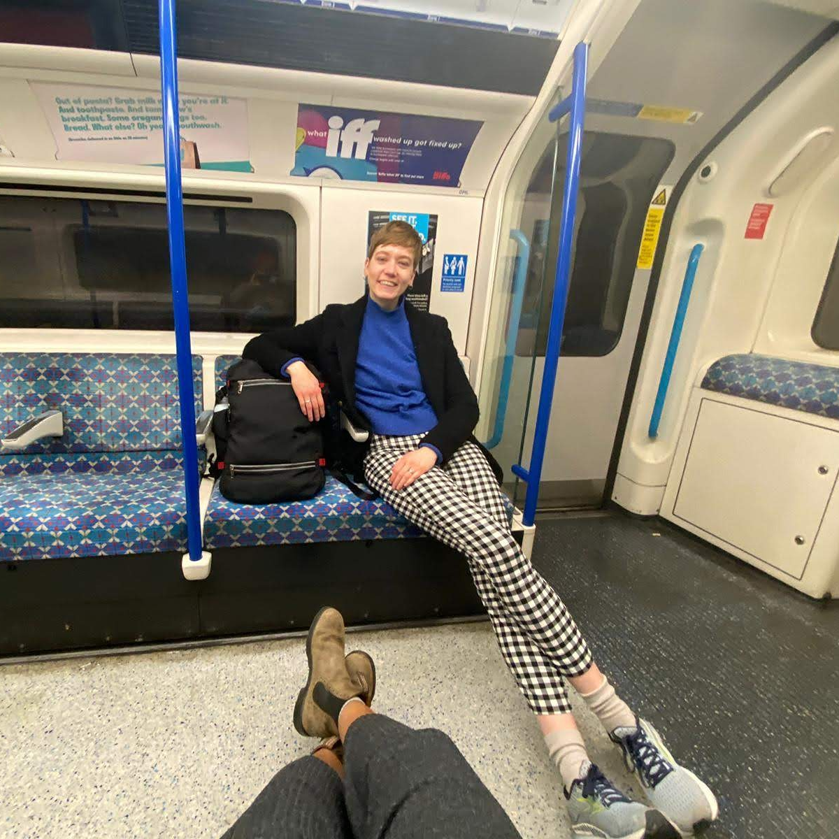 Roxy sits in an empty carriage of the London Undergrond, legs stretched out. They're wearing black and white checkered trousers, a blue jumper, and black coat. They lean on a big black bag.
