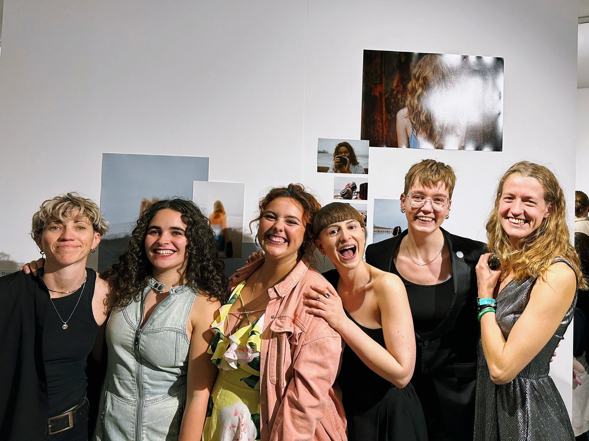 Six people are stood next to each other, all dressed up, posing in front of their photo exhibition wall at Photo Fringe Brighton in 2024