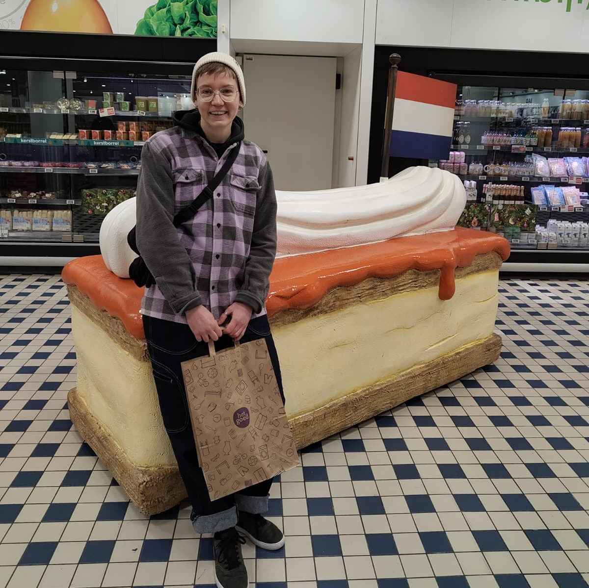 Roxy is wearing a thick winter jacket and a white hat, holding a brown paper bag. They're inside a Dutch shop, leaning on a giant plastic dessert called a 'tompoes' or 'tompouce'.