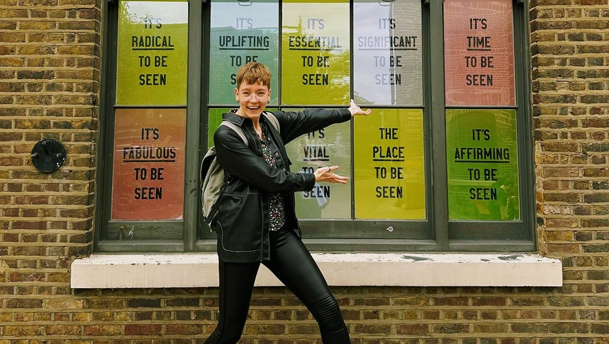 Roxy wears an all black outfit and is standing outside of the Queer Britain museum. They have a big smile on their face and point towards a window display that contains messages about queer visibility.