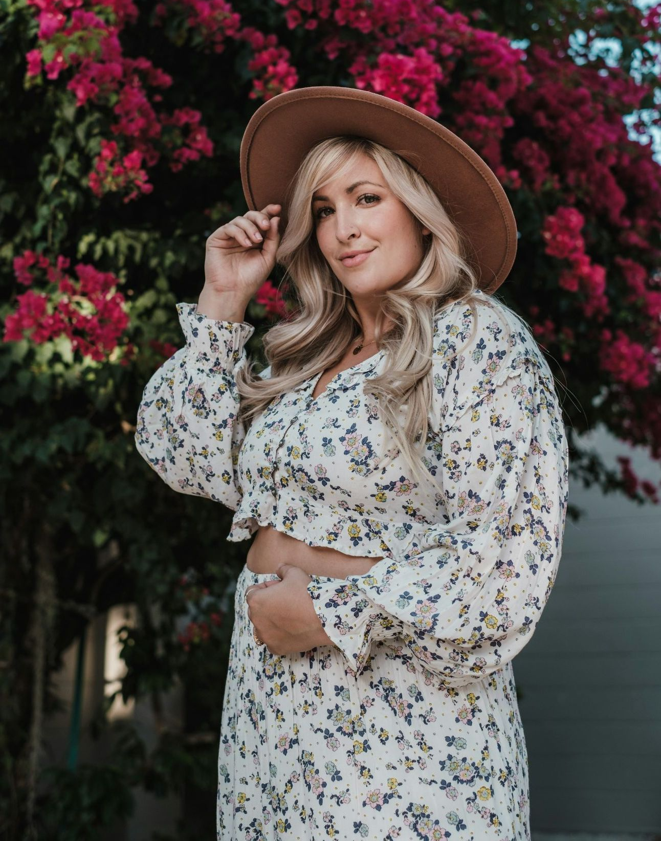 Woman with a curvy figure, light skin and blonde wavy hair, wearing a brown hat and floral co-ordinating top and bottoms in front of a bougainvillea plant