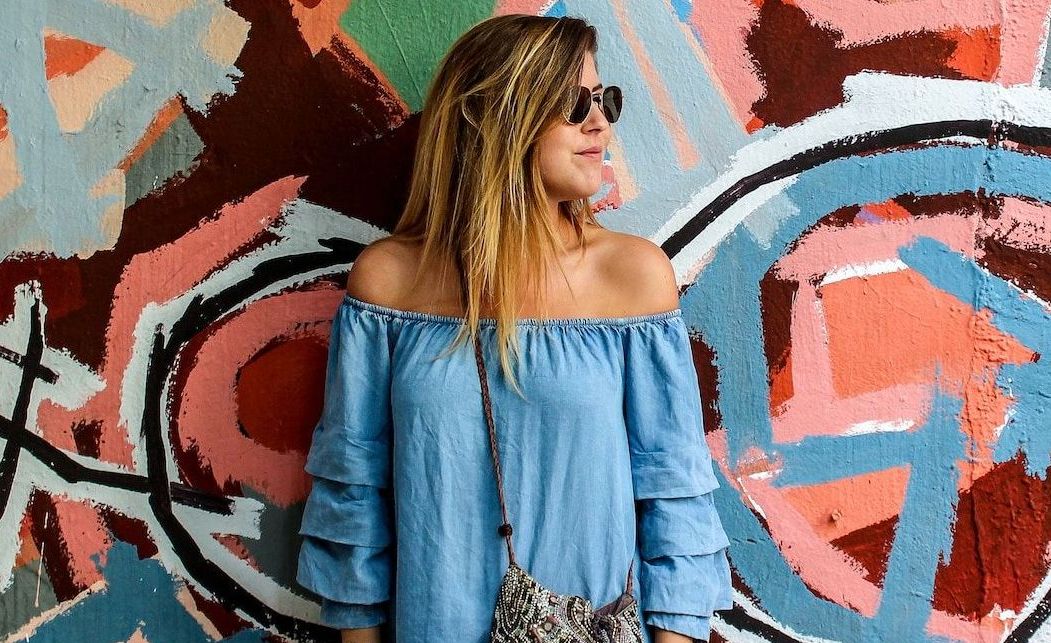 White woman with long blonde hair  wearing blue denim dress and cross-body bag standing in front of a colourful graffiti wall