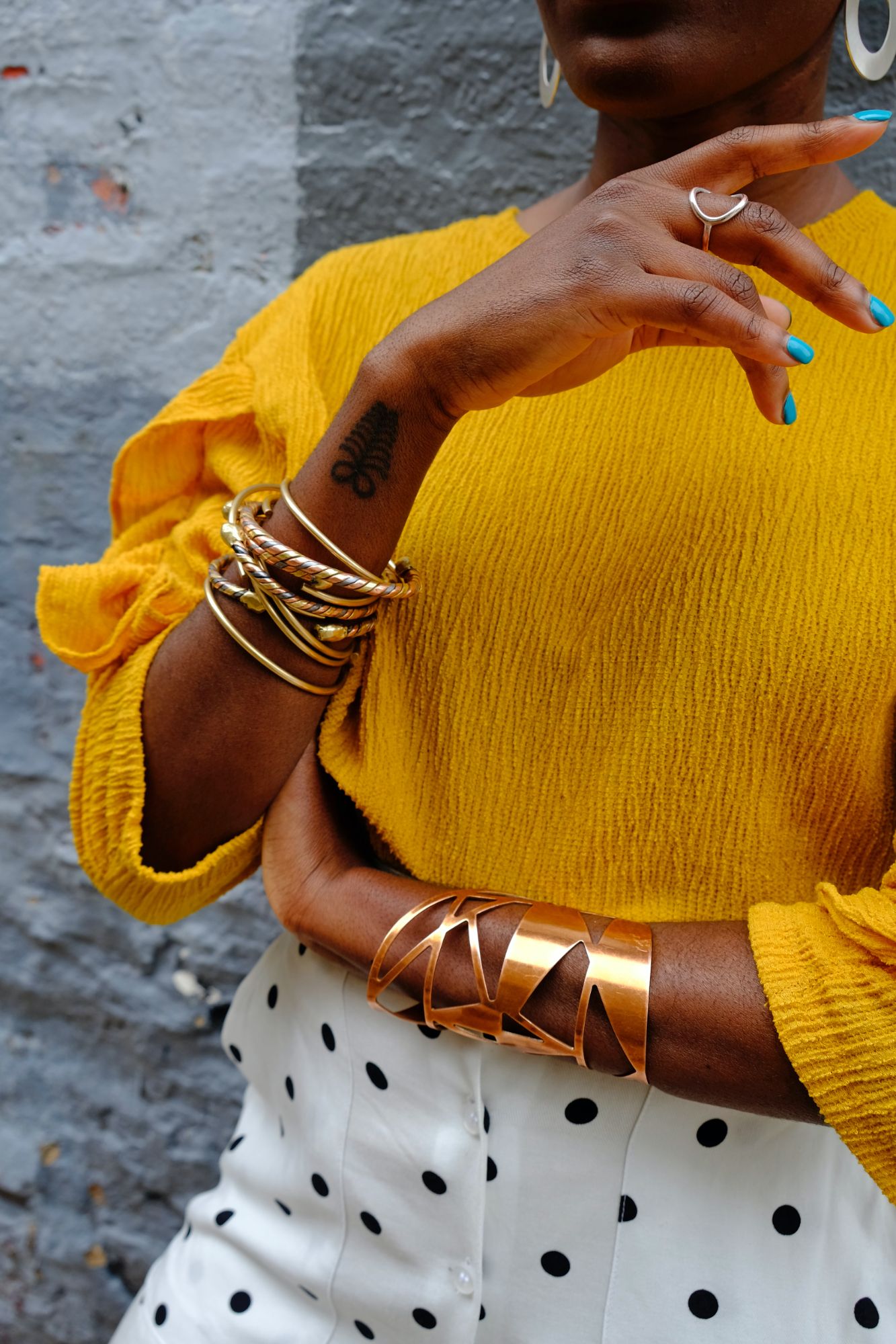 Stylish woman with black skin tone wearing gold bracelets, tattoo, mustard yellow top and spotty trousers