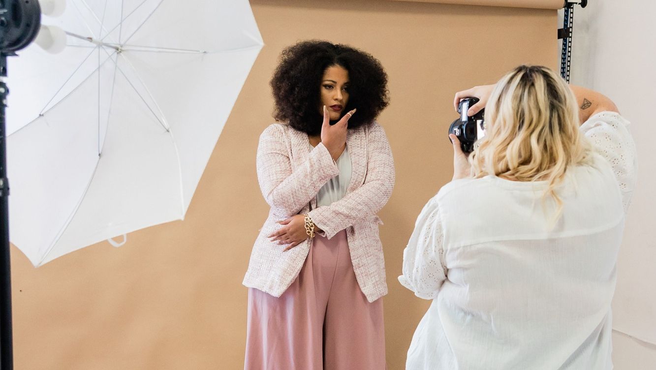 Female business owner in pale pink business casual clothes with afro hair posing in a photo studio having a photo shoot with a blonde female photographer in a white shirt