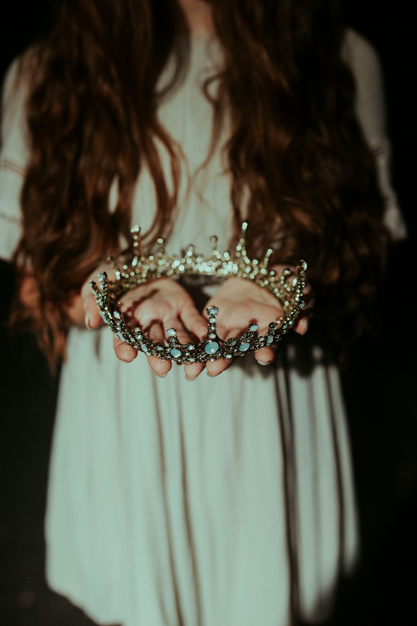 Princess in cream dress with brown wavy hair holding out a gold tiara