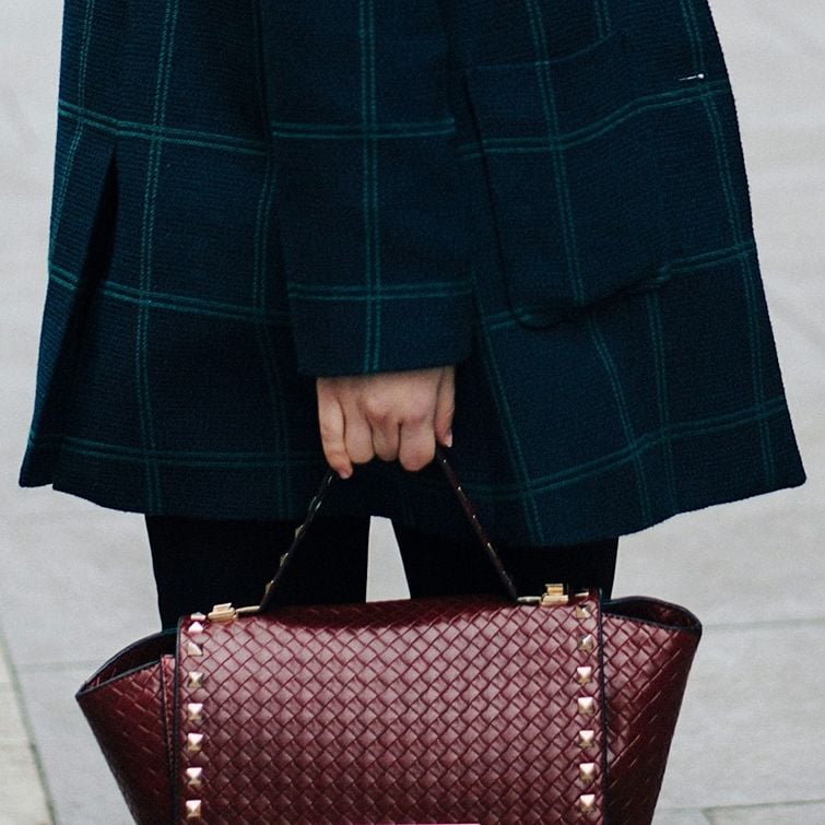 Close up of woman's arm in black checked blazer holding smart woven burgundy bag with gold studs