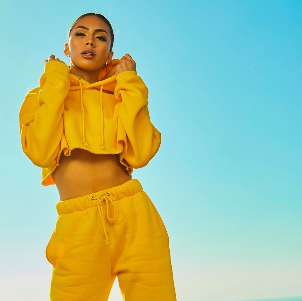 Dark skinned young woman with dark hair tied back wearing a yellow co-ord featuring cropped yellow hoodie and yellow jogging bottoms standing in front of a blue sky
