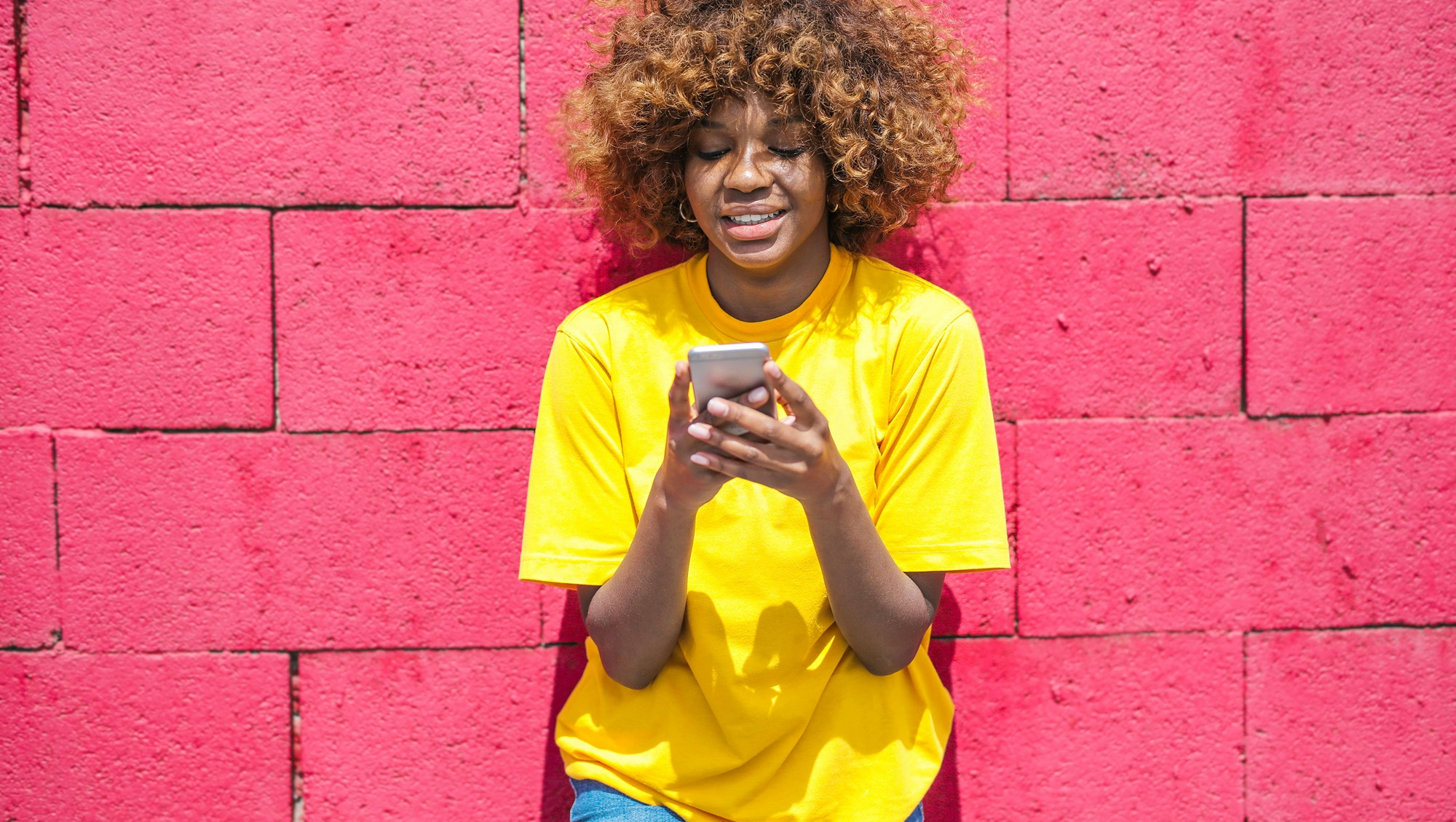 Dark skinned woman with loose afro curls in bright yellow T-shirt standing in front of a bright pink wall