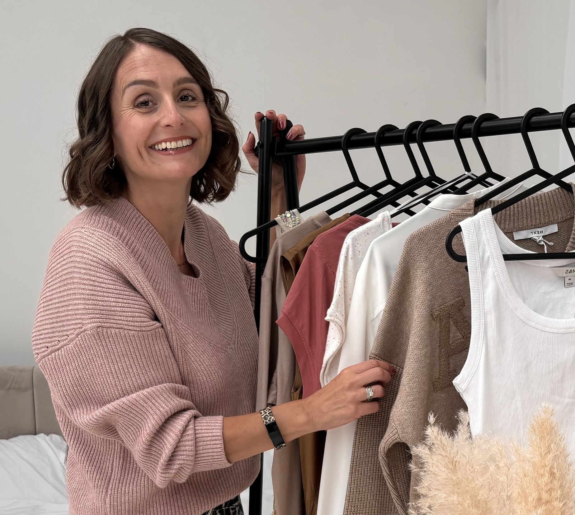 Personal stylist and colour analysis consultant in a light pink jumper and leopard print jeans touching a clothes rail with women's clothes in neutral colours hanging from it