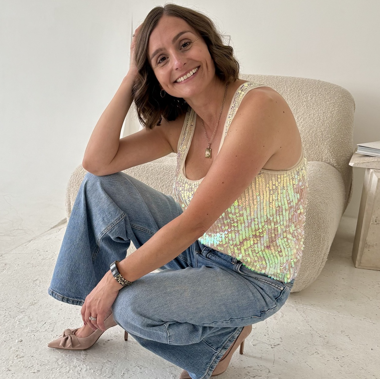 Personal stylist from Inspired Styling crouching dressed in cream sequinned vest and blue stonewashed jeans and taupe heels
