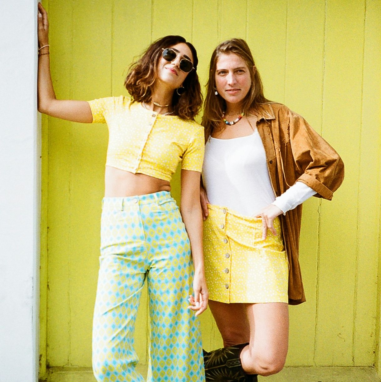 Two fair skinned women wearing butter yellow clothes, one with patterned pyjama pants, the other with a white vest and brown shirt and cowboy boots