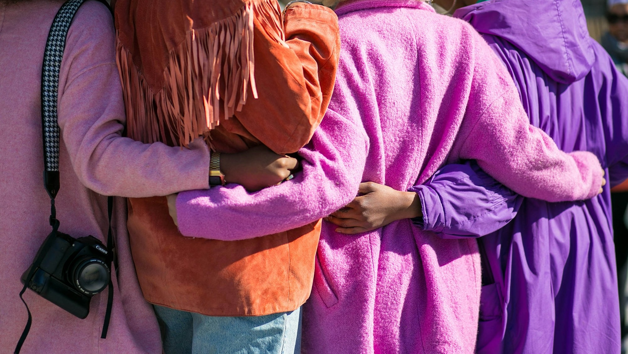 4 women with arms around them wearing colourful clothes in pink, coral and purple