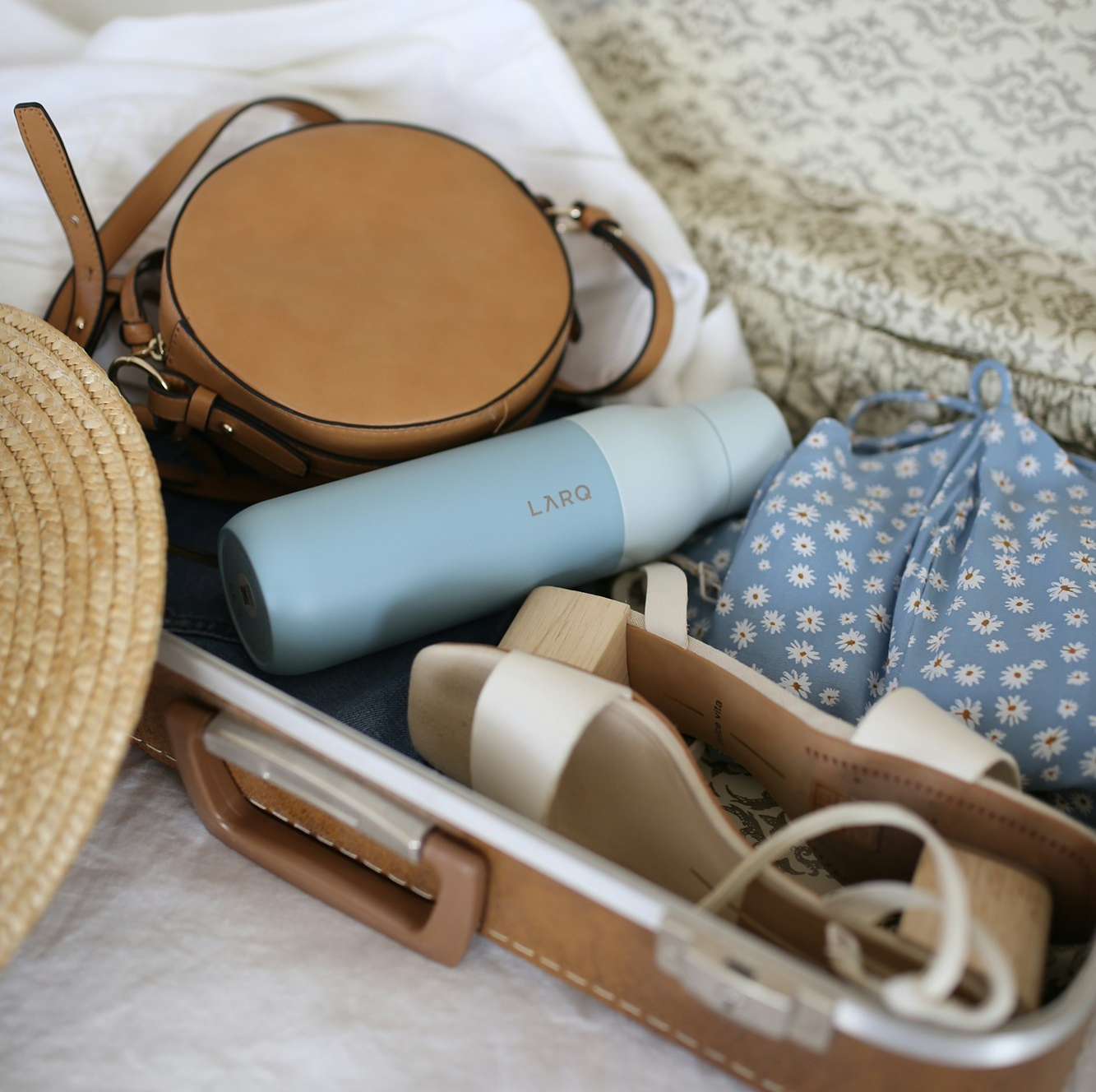 Brown retro leather suitcase open with tan handbag, blue water bottle, floral clothes and heeled sandals packed inside. 