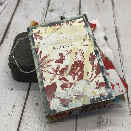 mini mixed media art journal with the word bloom on the cover is propped on a rusty tin