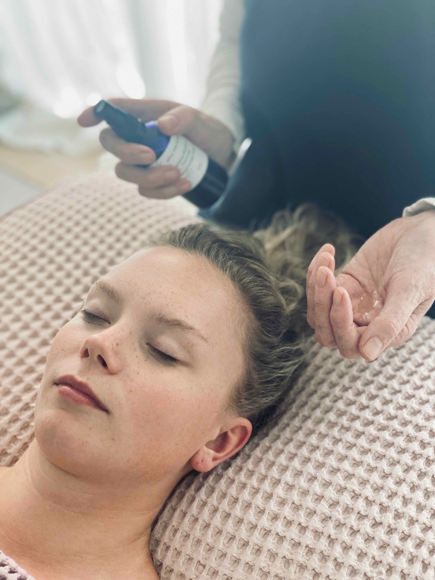 Lucy's hands holding handmade aromatherapy body oil, with a client relaxing on the therapy couch, enjoying a facial at Lucy's Facials.