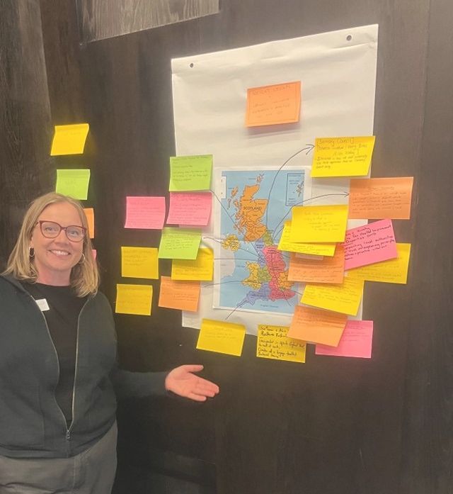 Cathy is smilng and pointing at a large map of the UK that has post it notes all over it.