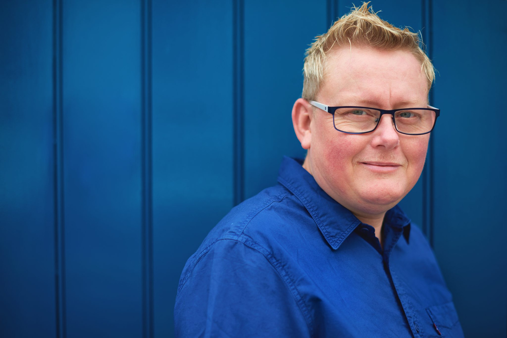Photo of Sarah Barnsley a white woman with short blonde hair and glasses in a blue shirt against a blue background