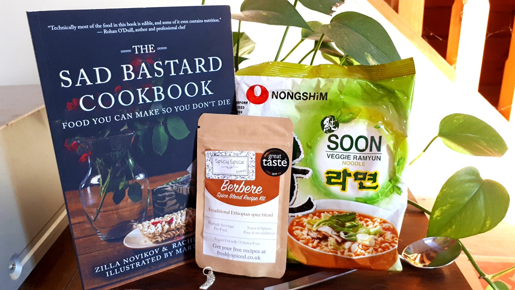 A photo of The Sad Bastard Cookbook next to a collection of goodies, including berbere spice mix, instant noodles, a spoon, and a silver pickle charm.