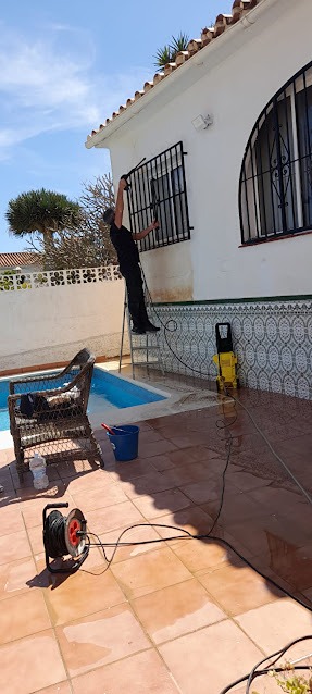 A maintenance worker using a high-pressure power washer to clean the exterior white walls of a villa near a swimming pool and terracotta patio area