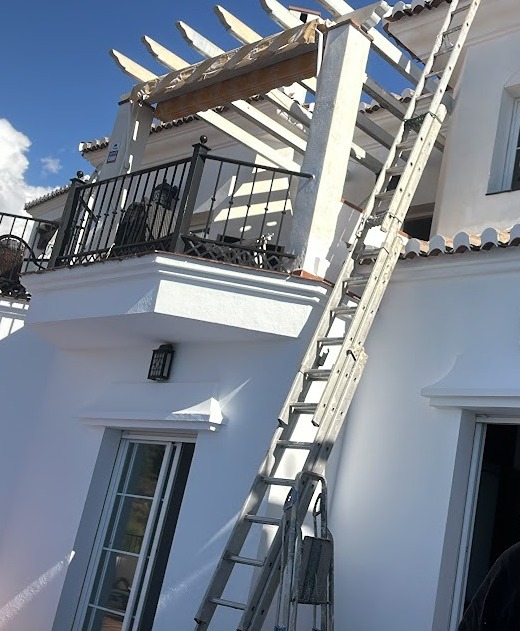 A professional window cleaning service in progress, featuring a tall aluminum ladder leaning against a white two-story villa with a black wrought-iron balcony under a bright blue sky