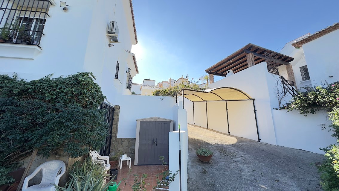A white-washed Spanish villa courtyard featuring lush green vines, outdoor furniture, and a curved metal carport structure under a clear blue sky