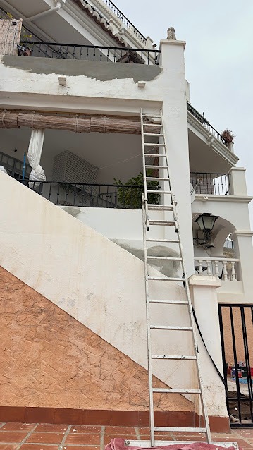 A tall aluminum ladder positioned against a white exterior wall next to a balcony with traditional wooden beam accents