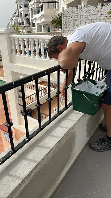 A close-up of a worker painting a black wrought-iron balcony railing on a white terrace with a green paint bucket