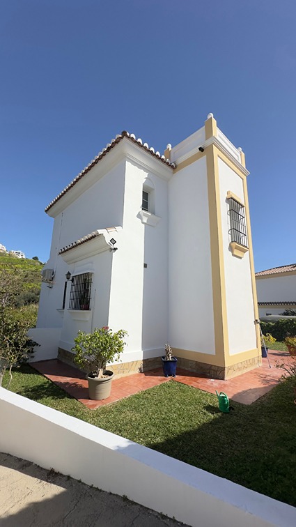 The exterior of a freshly painted white villa with yellow trim, traditional tiled roof, and a terracotta patio surrounded by a green lawn